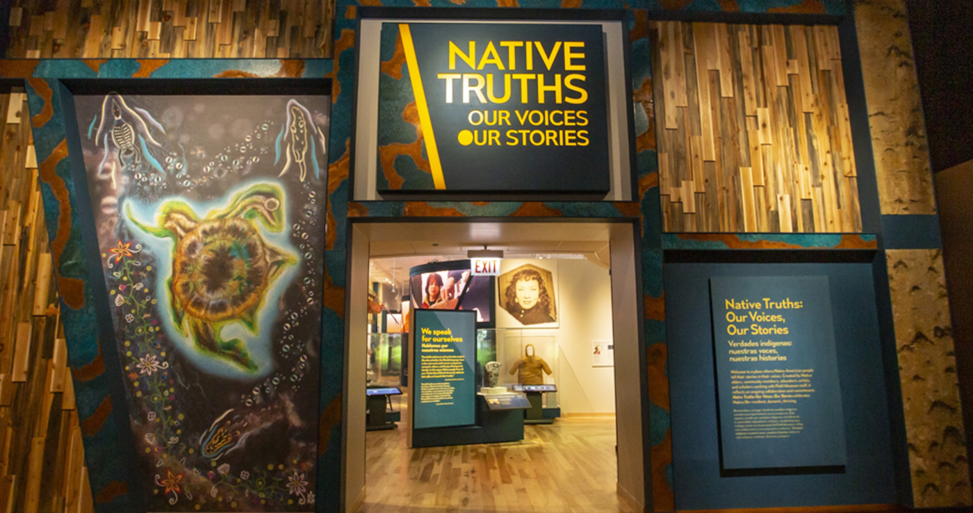 An entrance to the museum exhibition hall, with the title Native Truths: Our Voices, Our Stories above the doorway. An illustration by a Native artist is on the left of the doorway and a graphic panel to the right welcomes visitors.
