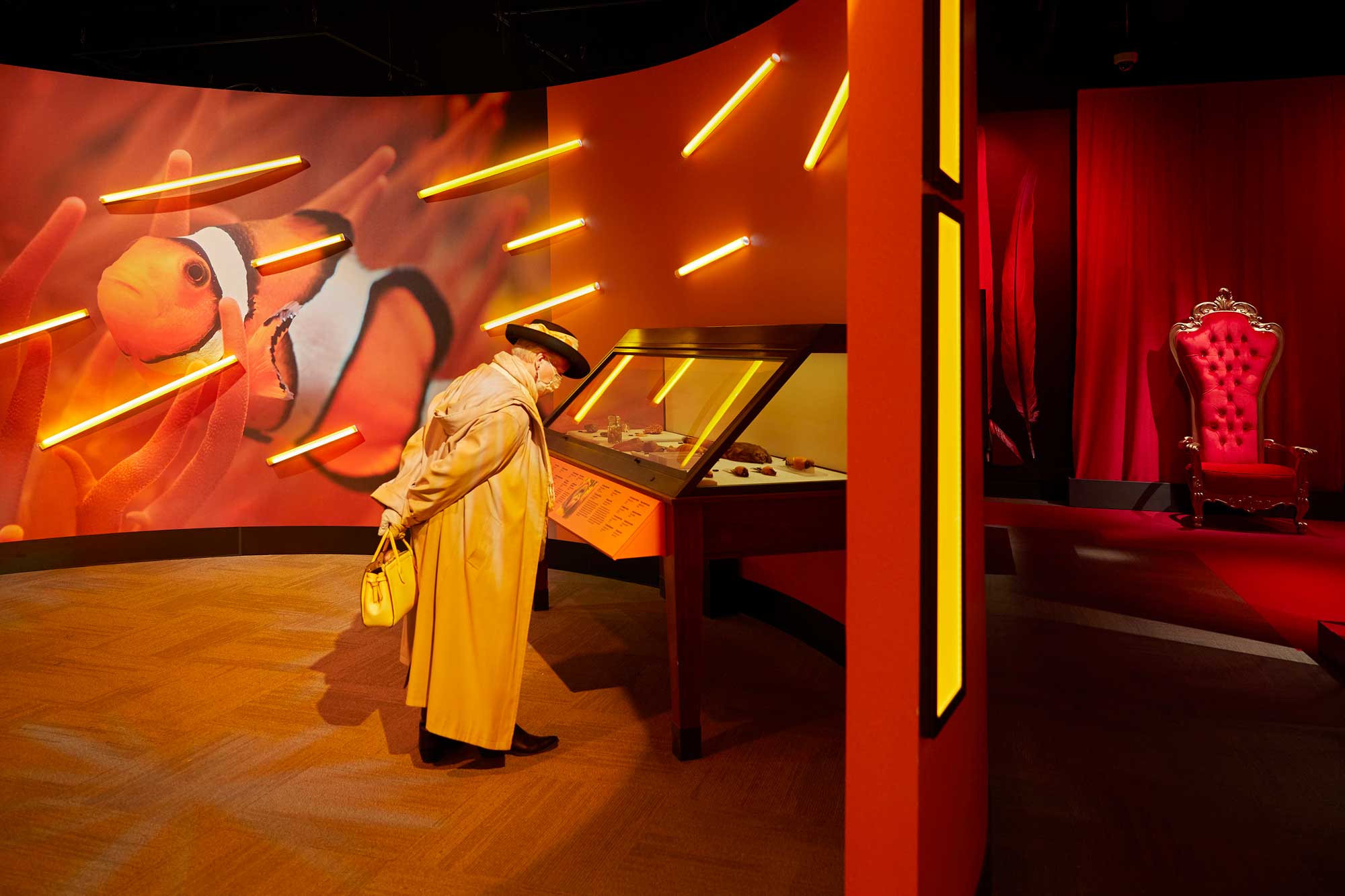 An adult reads a label on a display case in an orange gallery. The wall behind includes a giant clownfish mural and bars of orange light.