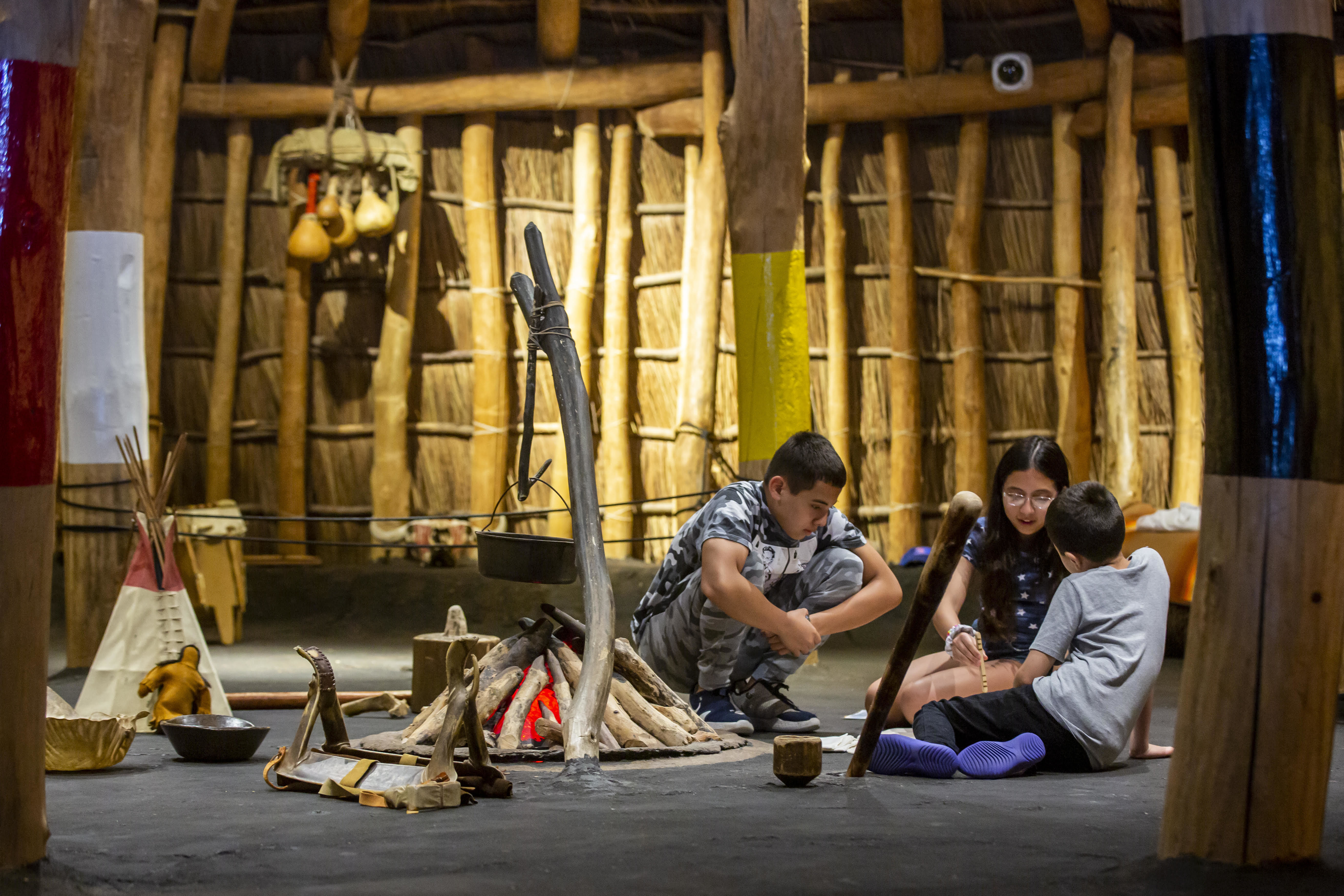 three children sit on the floor of the Pawnee Earth Lodge, interacting with each other and replicas of items from the 1800s.