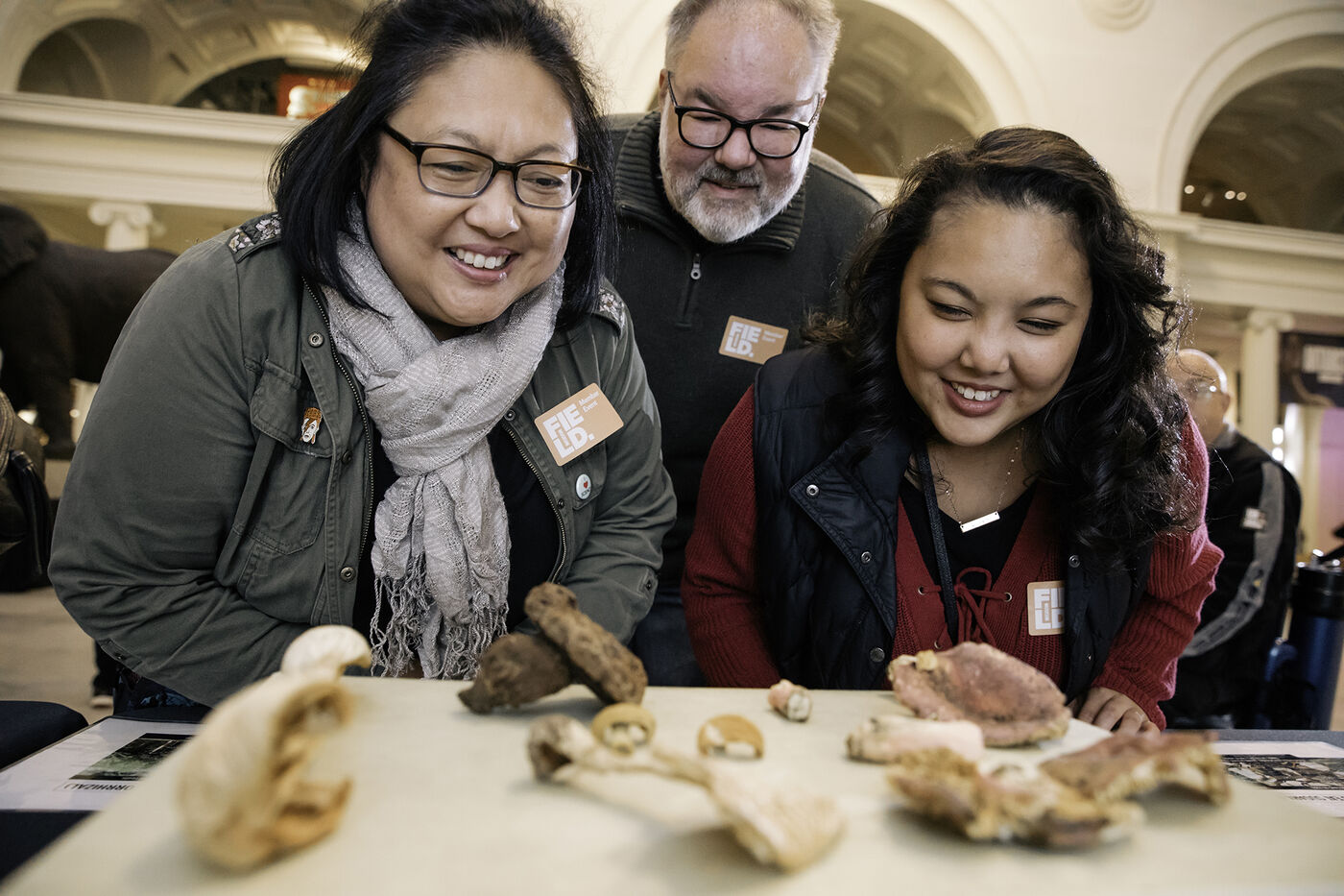 A group of three Field Museum members gather in Stanley Field Hall during an event. They are leaning forward and smiling as they take a closer look at specimens arranged on a table.