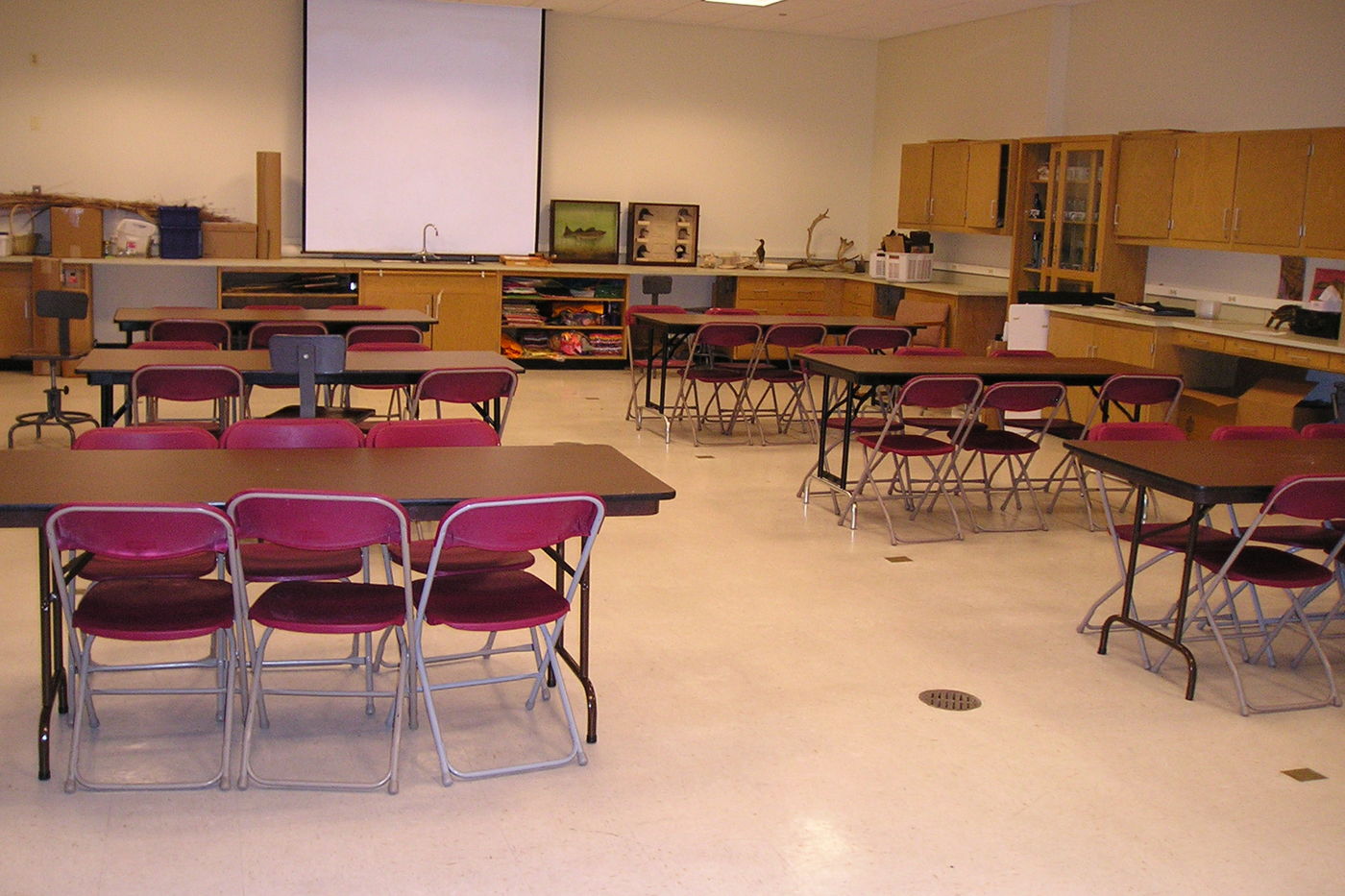 A view of Classroom B. Six desks are arranged in two rows, facing a projector on the wall in the front of the room. Each table seats six guests. Counters and cabinets line the walls of the room.