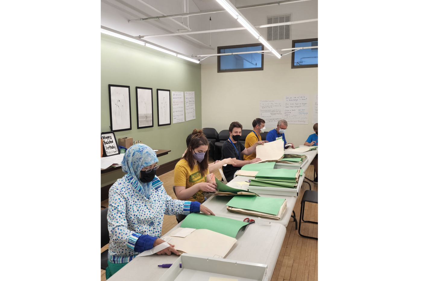 Several people sit along tables stacked with file folders of dried plant material, museum herbarium specimens.