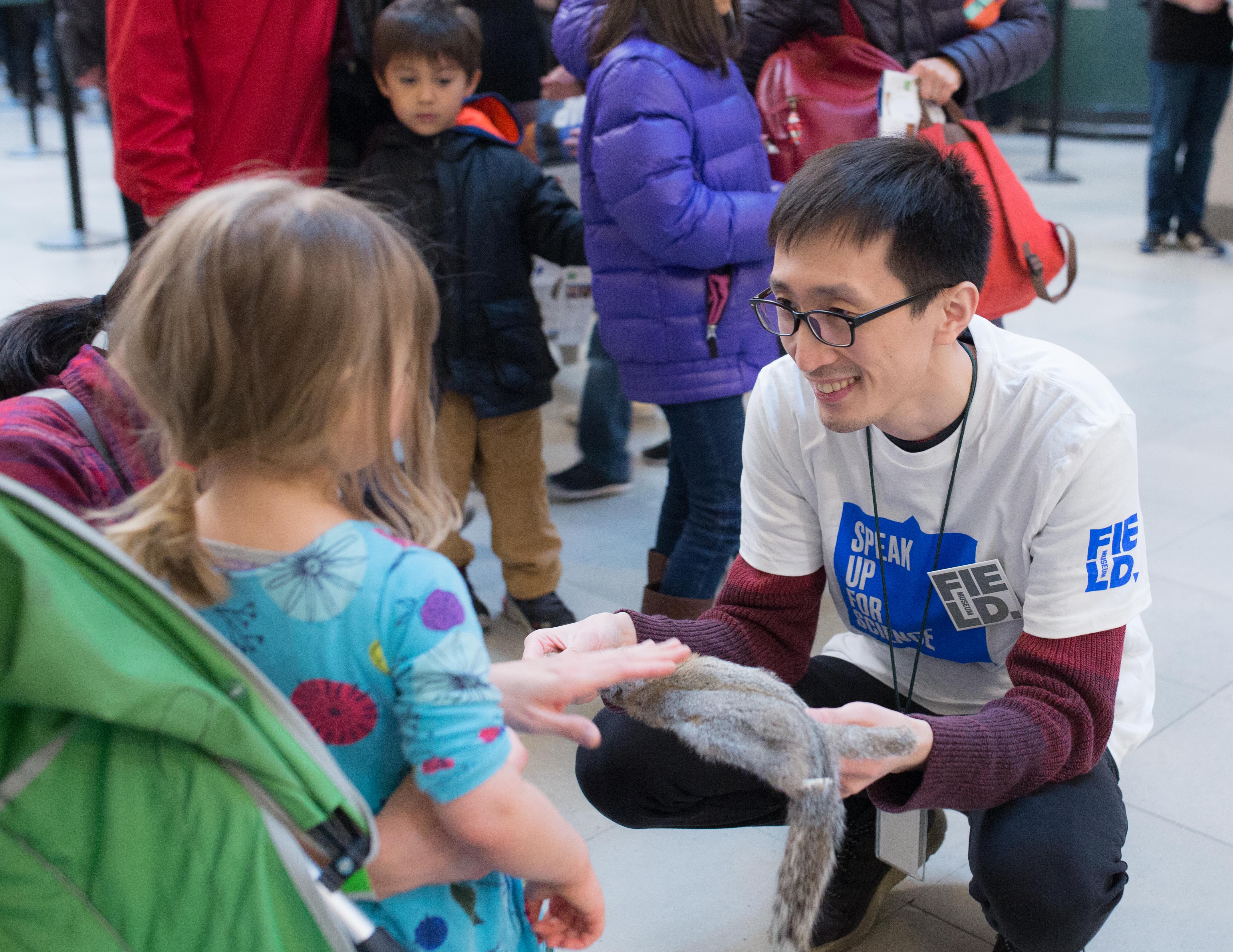 A museum staff member stoops down to interact with a young visitor, offering a furry specimen for the visitors to touch.