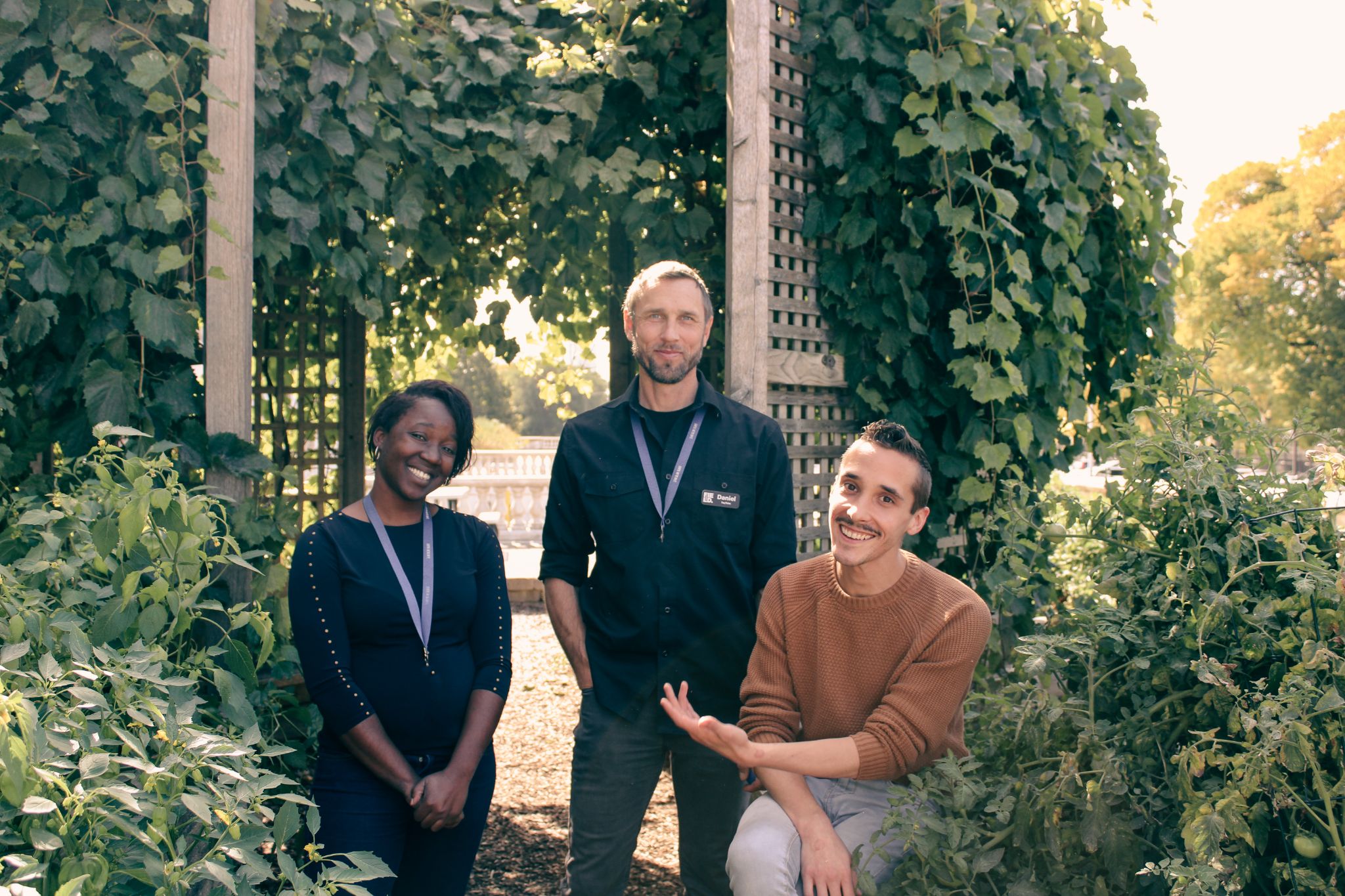 Three people in the Museums garden, one standing between two seated on the edge of raised garden beds.