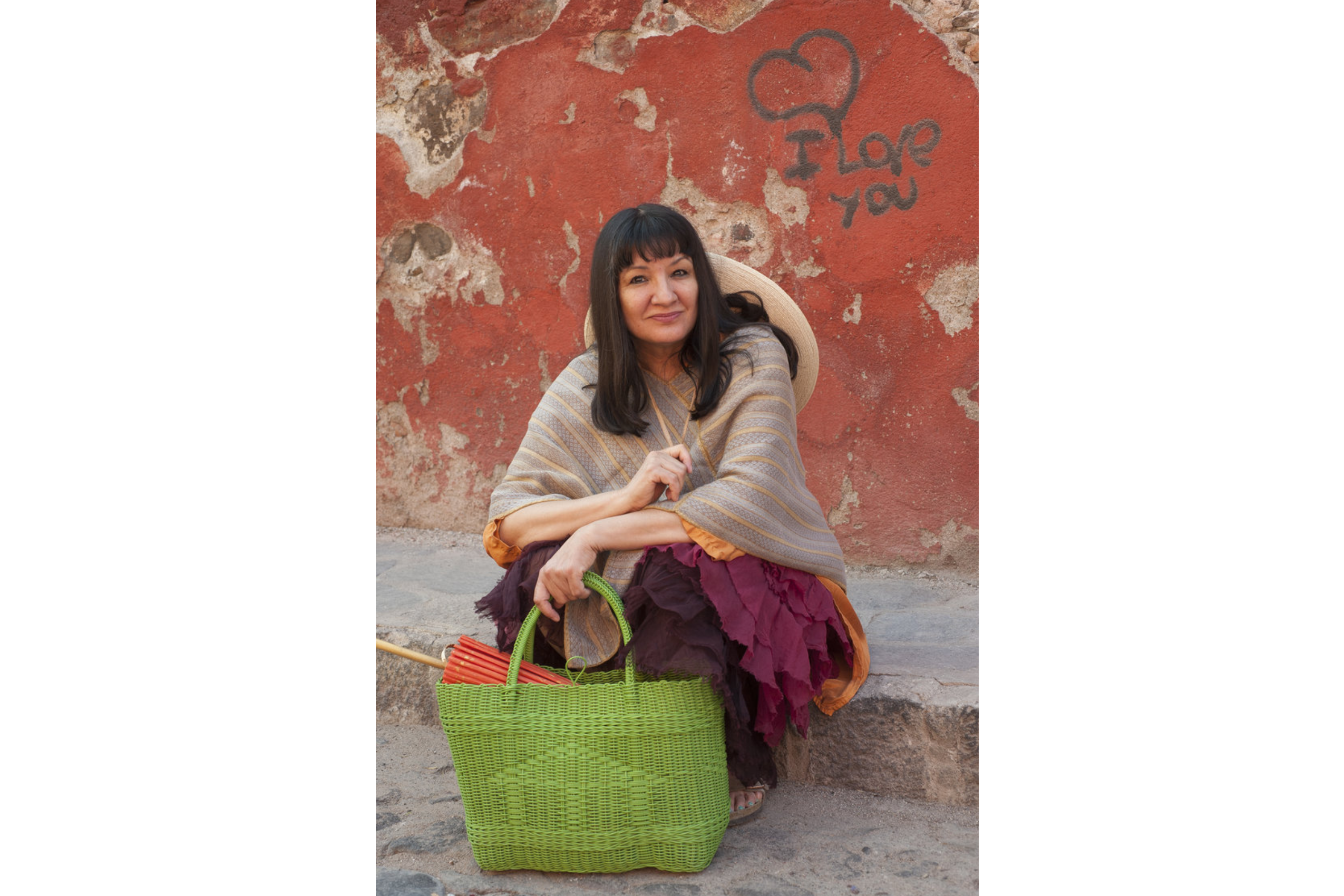 A woman sits on a curb, a green woven bag sits on the ground in front of her. The orange wall of the building behind her had "I love you" written in grey.