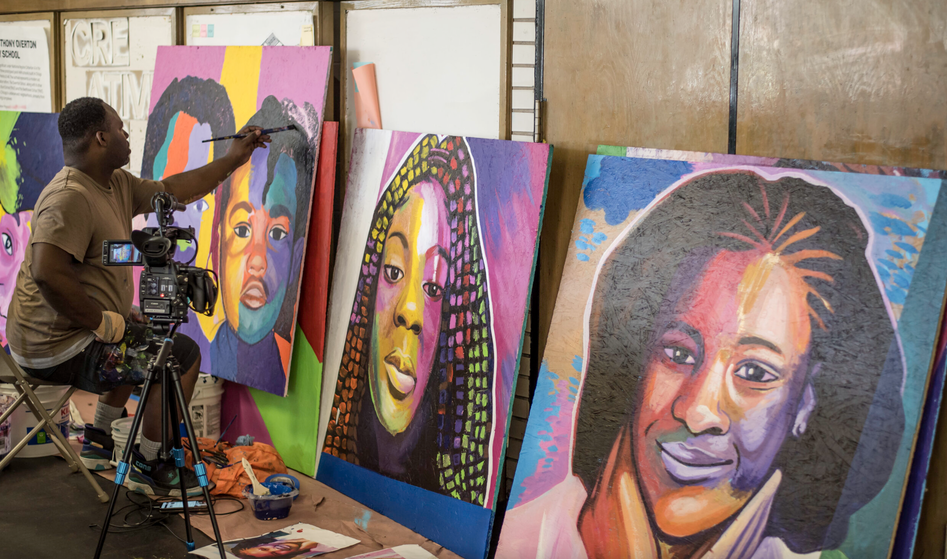 A African American man seated on a stool, painting a large canvas with brightly colored portraits of African American girls and  women. Two other portraits are propped up next to the one he is working on.