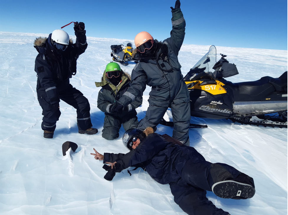 Four researchers, one lying on the snow covered ground, pose with a large black meteorite. Two snowmobiles can be seen in a completely snow covered landscape.