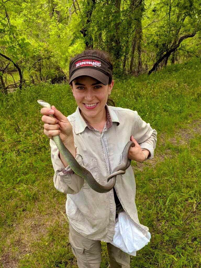 A woman stands in a grassy area with shrubbery behind her as she holds a snake with both hands.