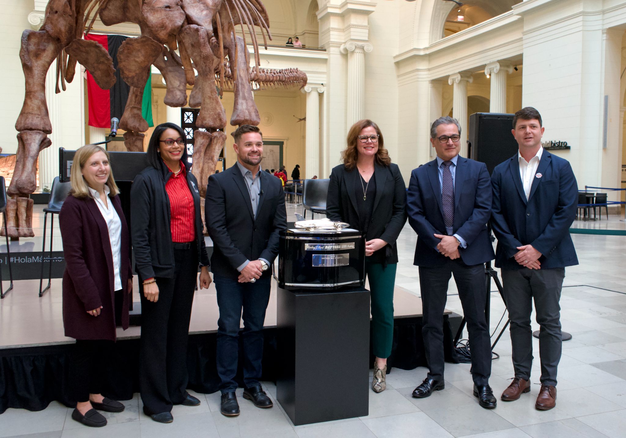Six people stand beside a podium with a dinosaur fossil behind them.