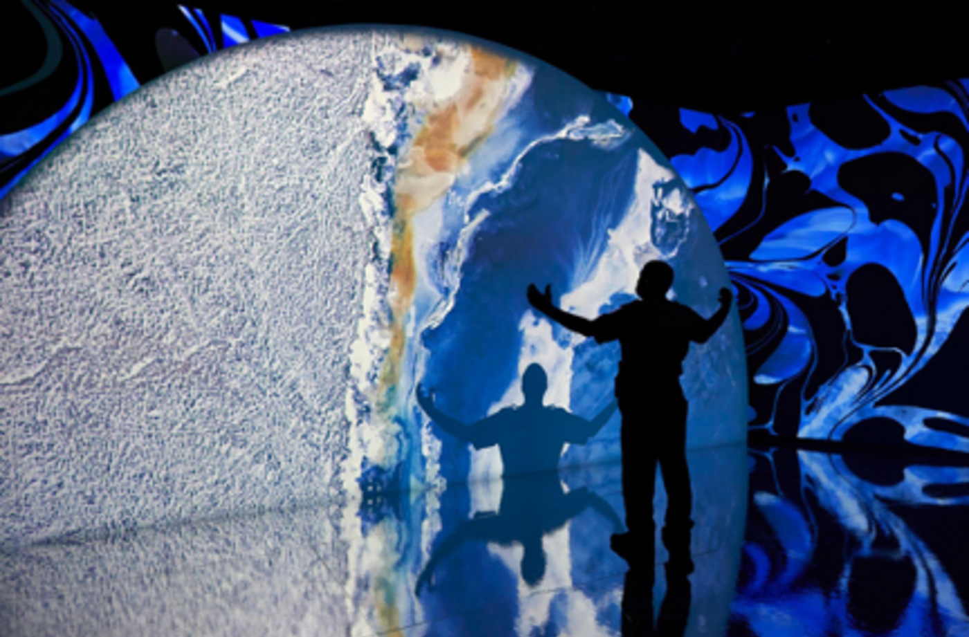 A person stands in a multimedia exhibition space with his arms outstretched. He's seen in silouhette, casting a shadow on a wall with an arial view of earth being projected.