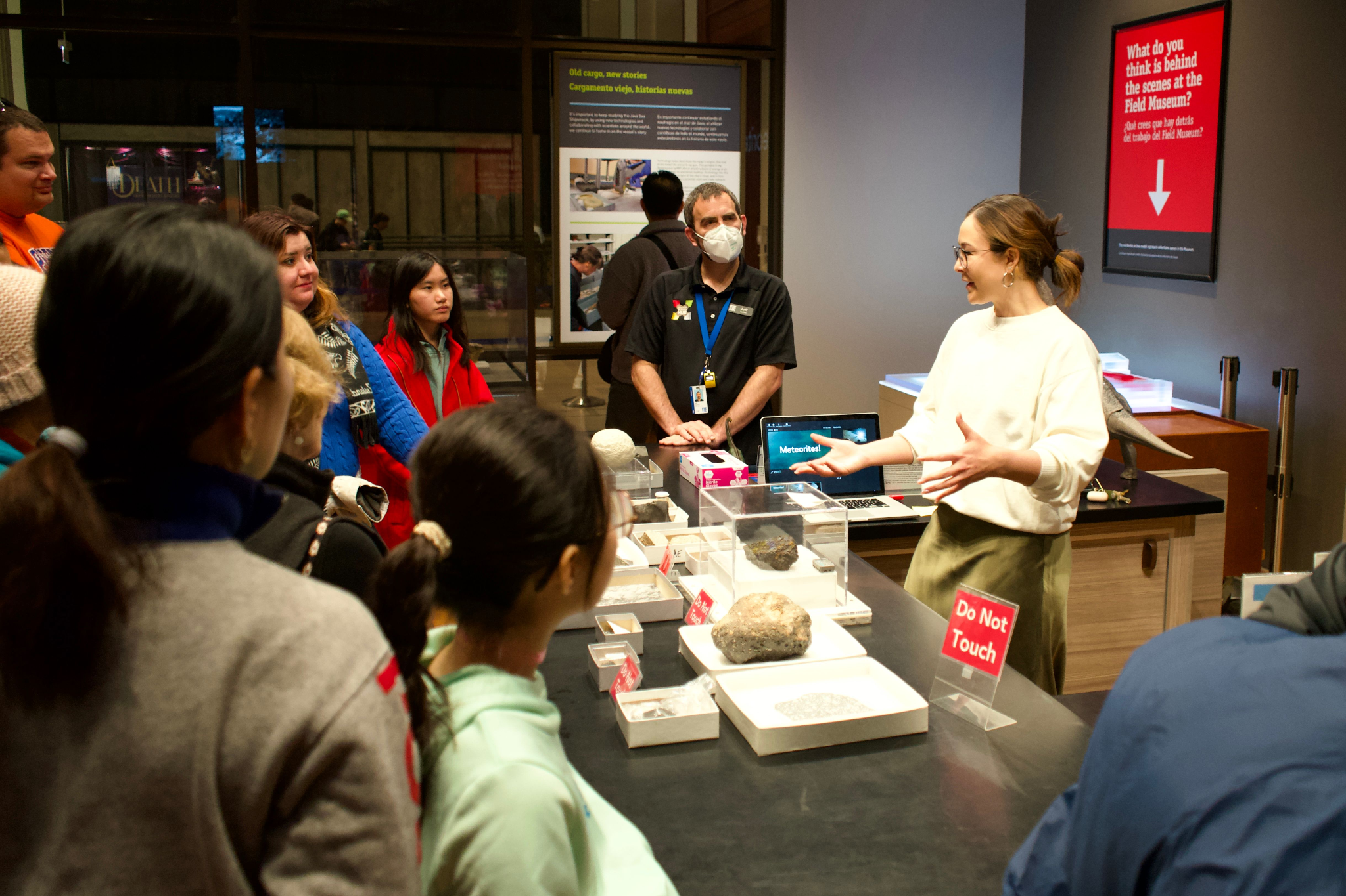 A woman stands engaging with a group of people across a table that is covered with museum specimens. A man stand looking on in the background