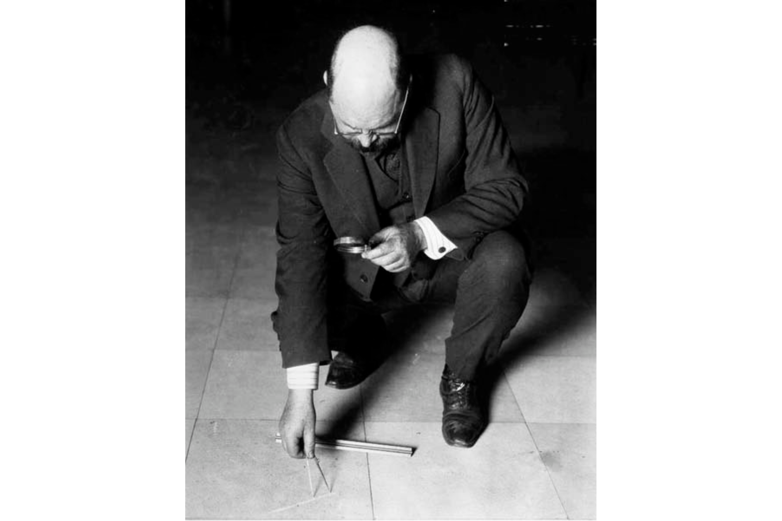 A man wearing a dark colored suit stoops down holding a magnifying glass and pointing at the floor