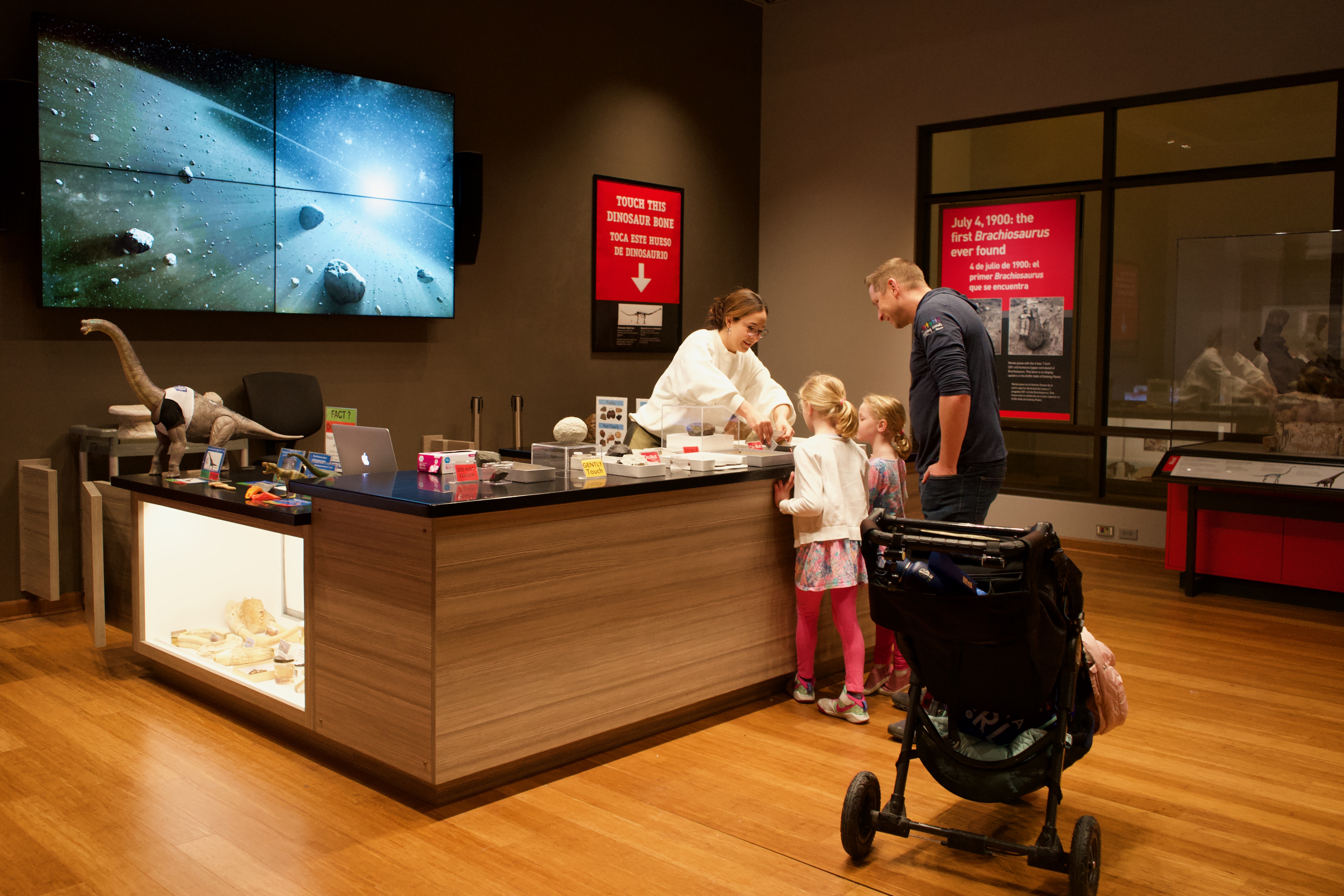 An adult and two children interact with a museum staff member who stands behind a counter in the Science Hub. On the counter are objects and specimens related to the content in the exhibit. A large video screen hangs on the wall behind the counter.