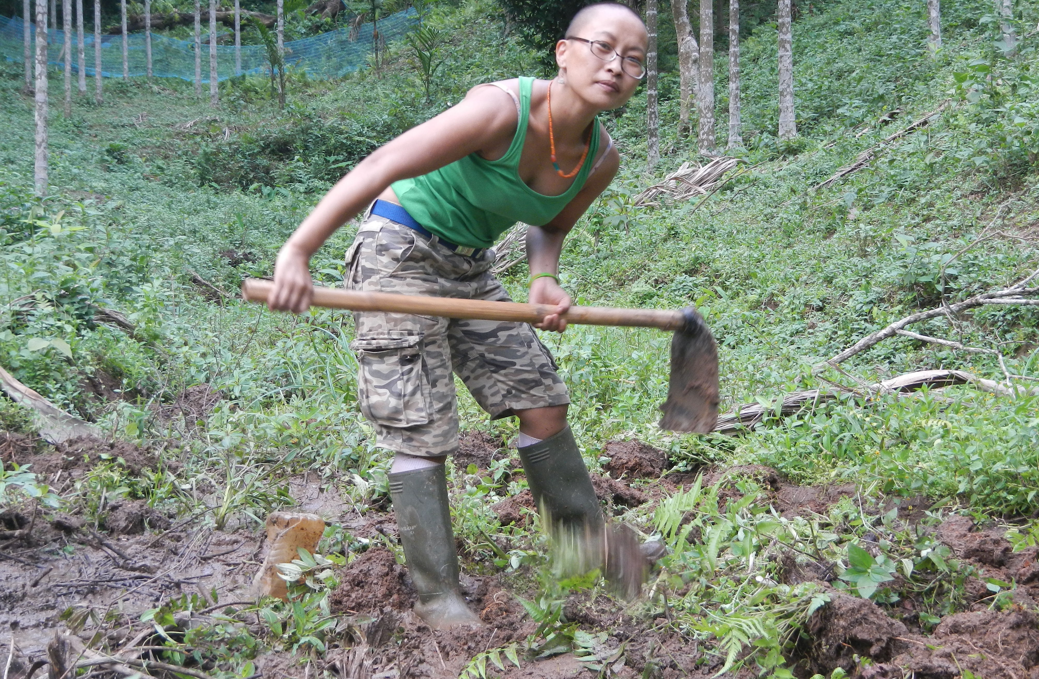 A women in a green tank top and boots, holding a garden hoe