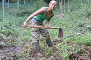 A women in a green tank top and boots, holding a garden hoe