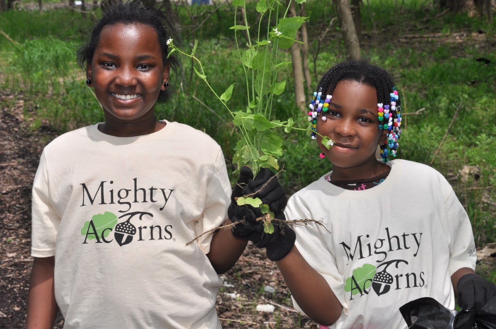 Two students wearing Mighty Acorns t-shirts hold up garlic mustard