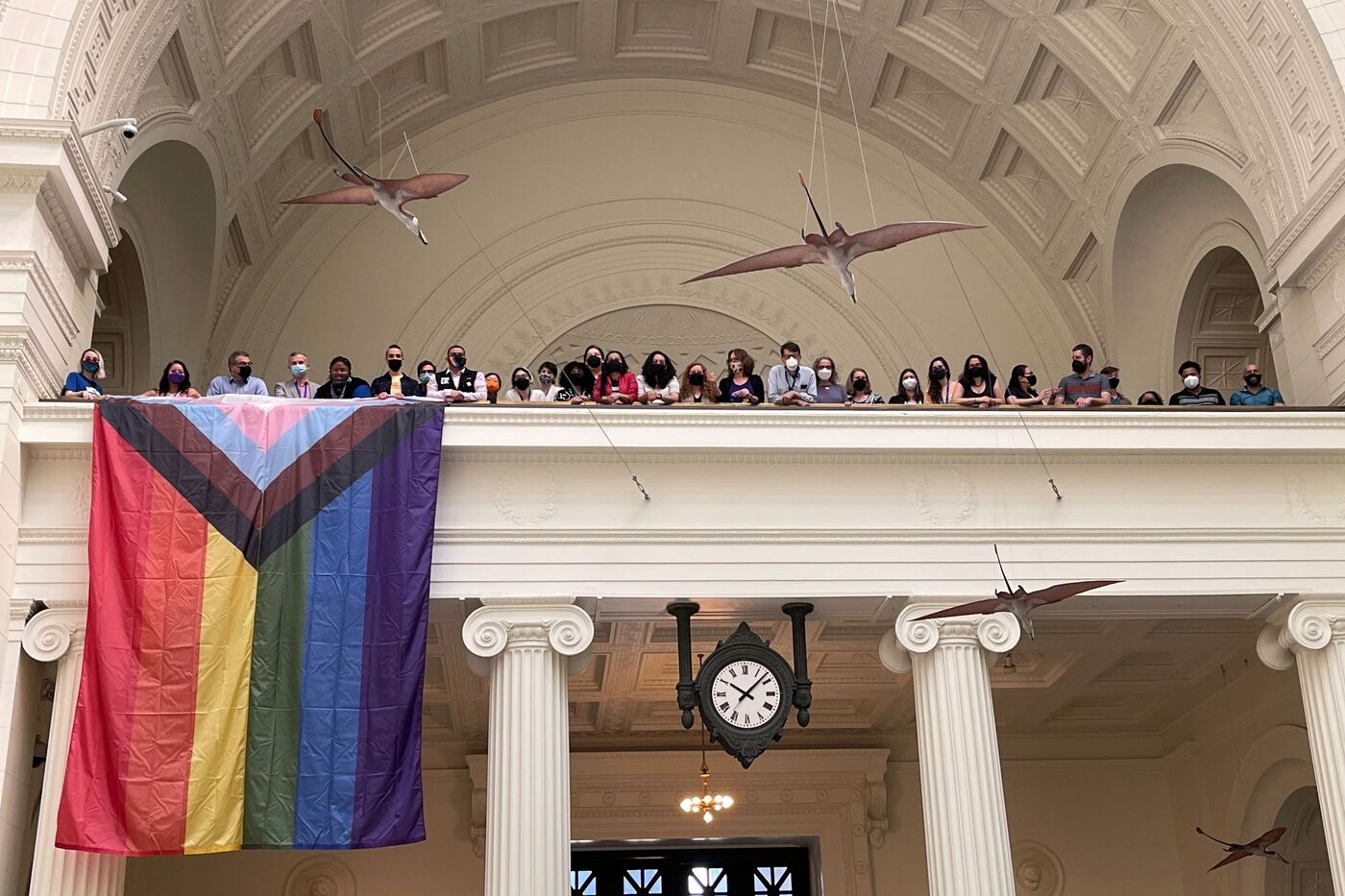 A group of people like the mezzanine of the museum, with the progressive pride flag hanging.