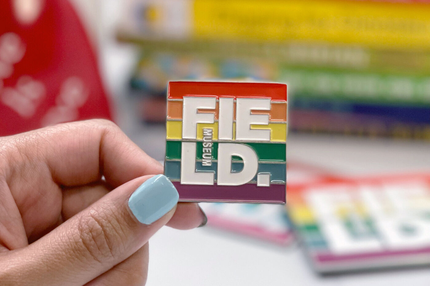 A person holding a square enamal pin. The pin is square, with a rainbow background and the Field Museum logo in white.