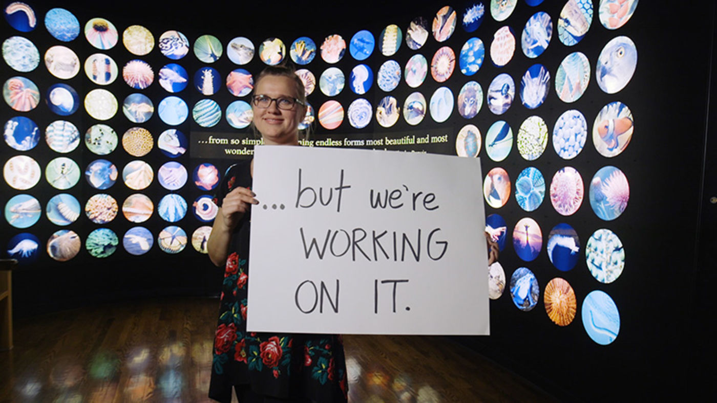 Woman holding a handwritten sign, standing in front of a mural of images from nature.