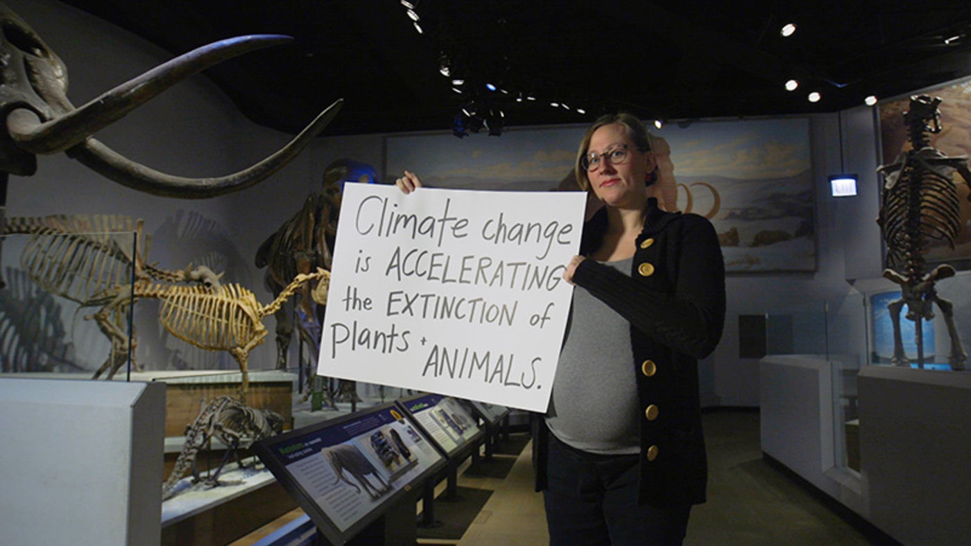 Woman holding a handwritten sign, standing in a museum gallery of large animal skeletons, including one with large tusks
