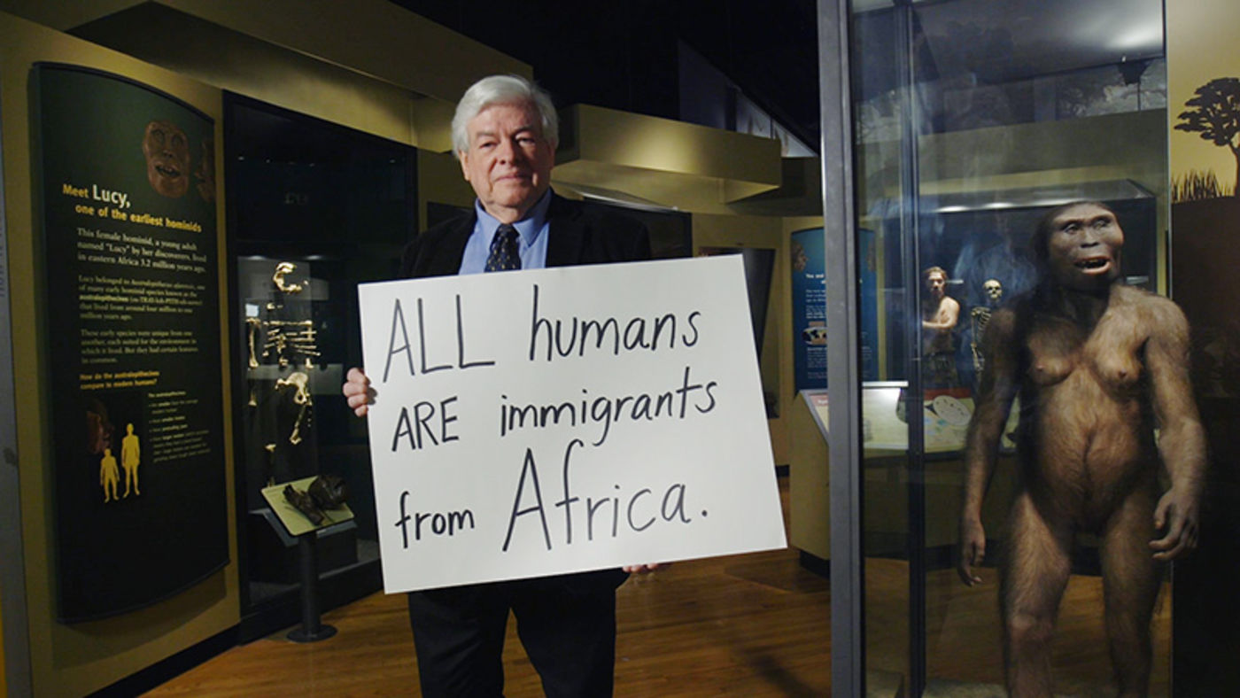 Man holding a handwritten sign, standing next to a figure of an early human relative.