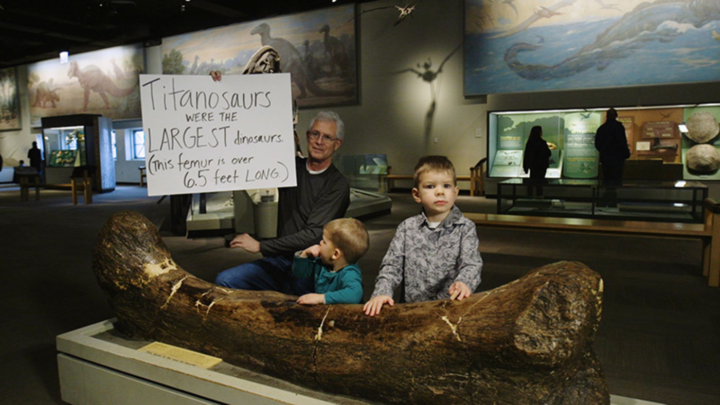 A man holding a handwritten sign, alongside two young boys, all behind a large dinosaur leg bone
