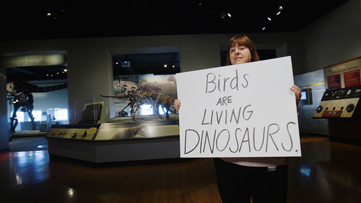 A woman holding a handwritten sign in front of a large museum display of animal skeletons.