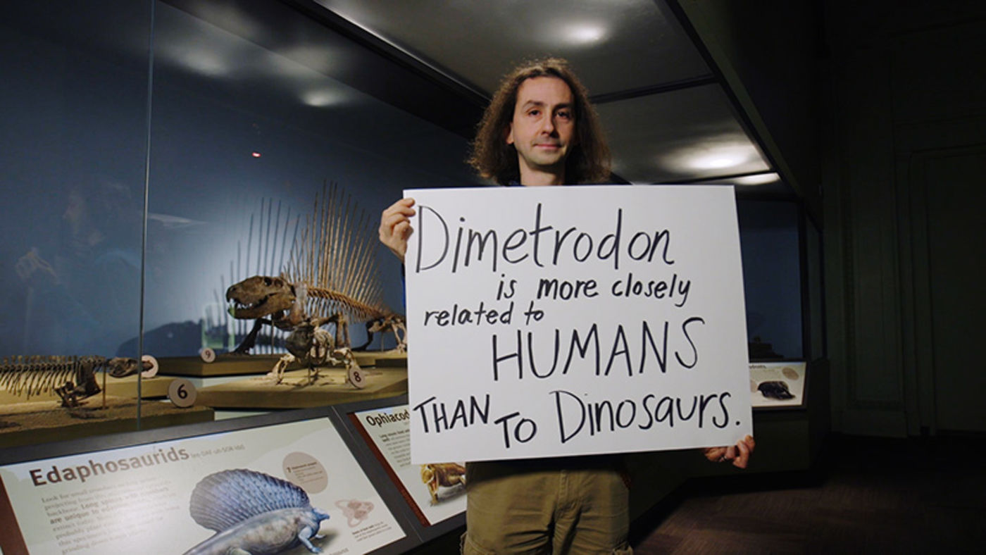 A man holding a handwritten sign, standing in front of a museum case containing a fossil animal with many spikes down its spine