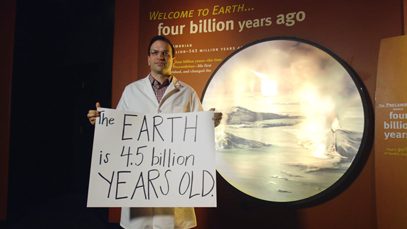 A man in a white lab coat holding a handwritten sign and standing in front of a museum exhibit showing volcanoes erupting.