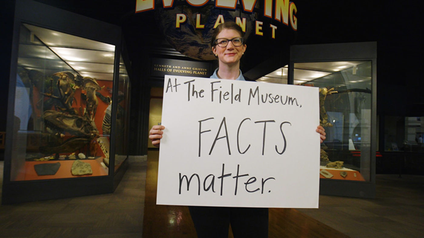 A woman holding a poster with a handwritten statement on it, standing in front of museum cases with fossils and other objects. 