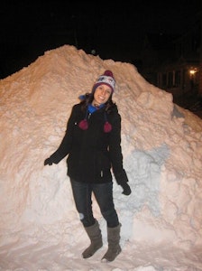 A smiling woman in a winter hat, gloves, and coat stands in front of a massive pile of snow