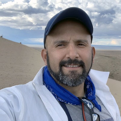 A man in a baseball hat and white coat stands in front of sandy hills and white clouds