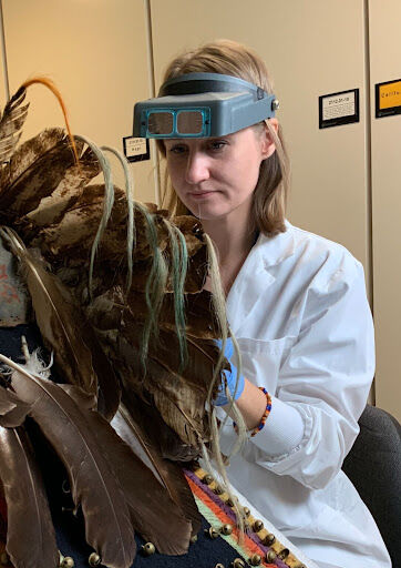 A woman wearing magnifying-goggles and a white lab coat works on an ornate feathered object