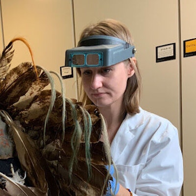 A woman wearing magnifying-goggles and a white lab coat works on an ornate feathered object
