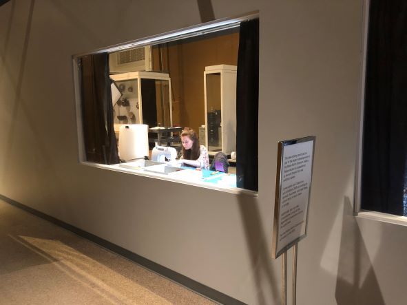 A woman works at a machine behind a glass window in the wall of a museum gallery.