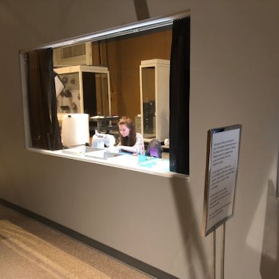 A woman works at a machine behind a glass window in the wall of a museum gallery.