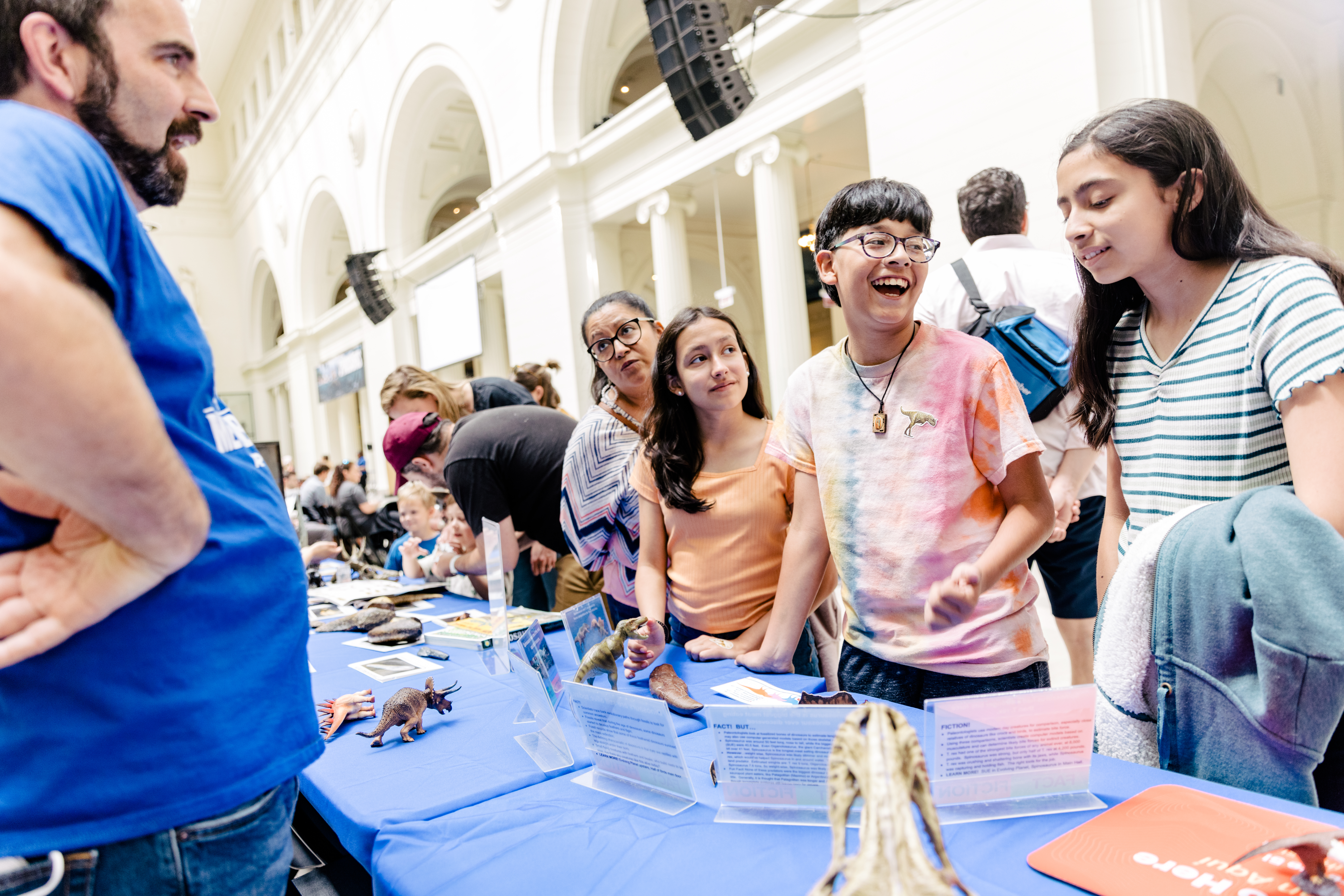 Several museum visitors stand at a table engaging with a staff member, who stands on the other side of the table.