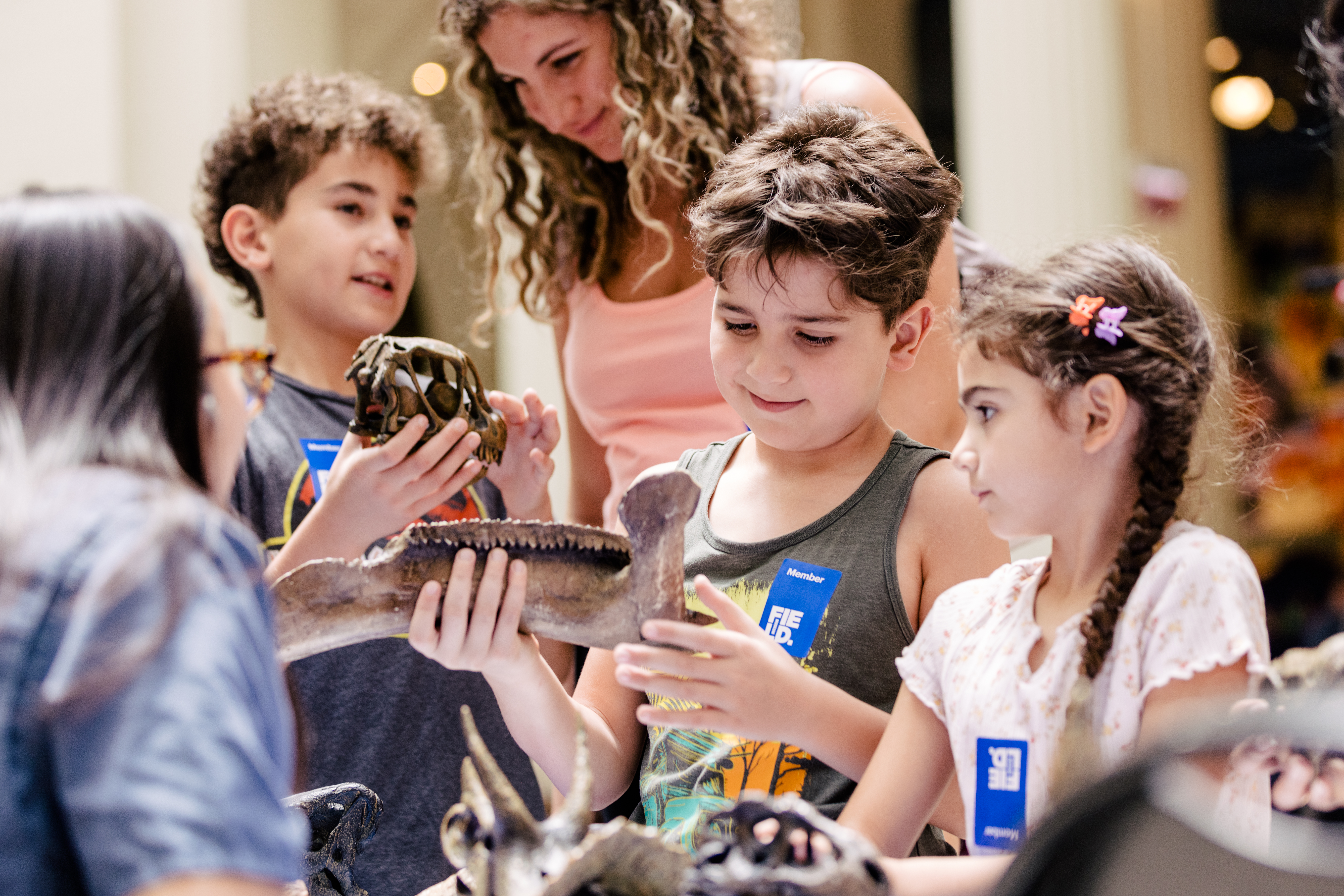 A boy looks down at a fossil bone he is holding as a young girl stands next to him looking on. Behind them are an adult and another boy holding a fossil skull.