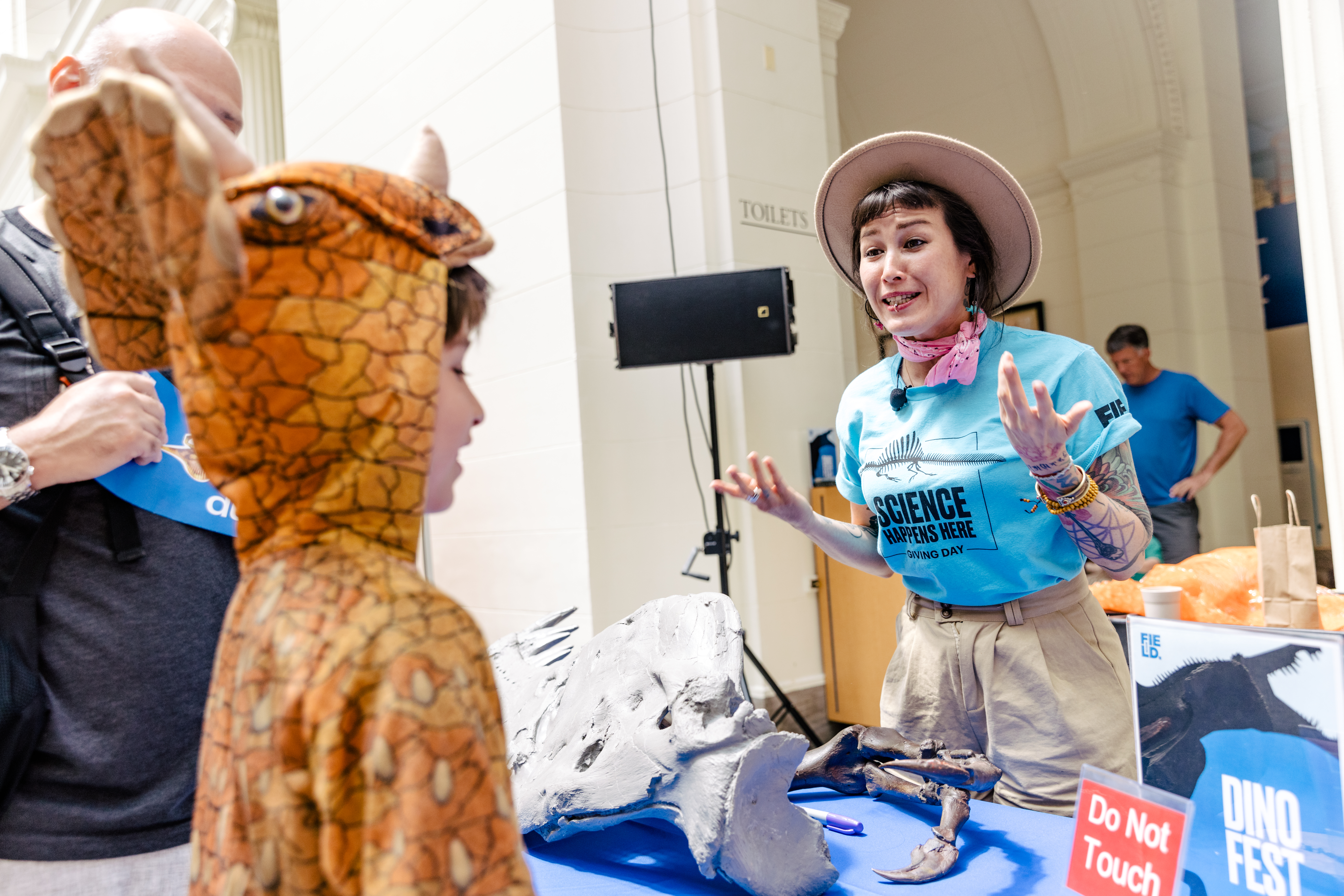 a person wearing a hat and bandana around their neck gestures and talks to a child in a dinosaur costume