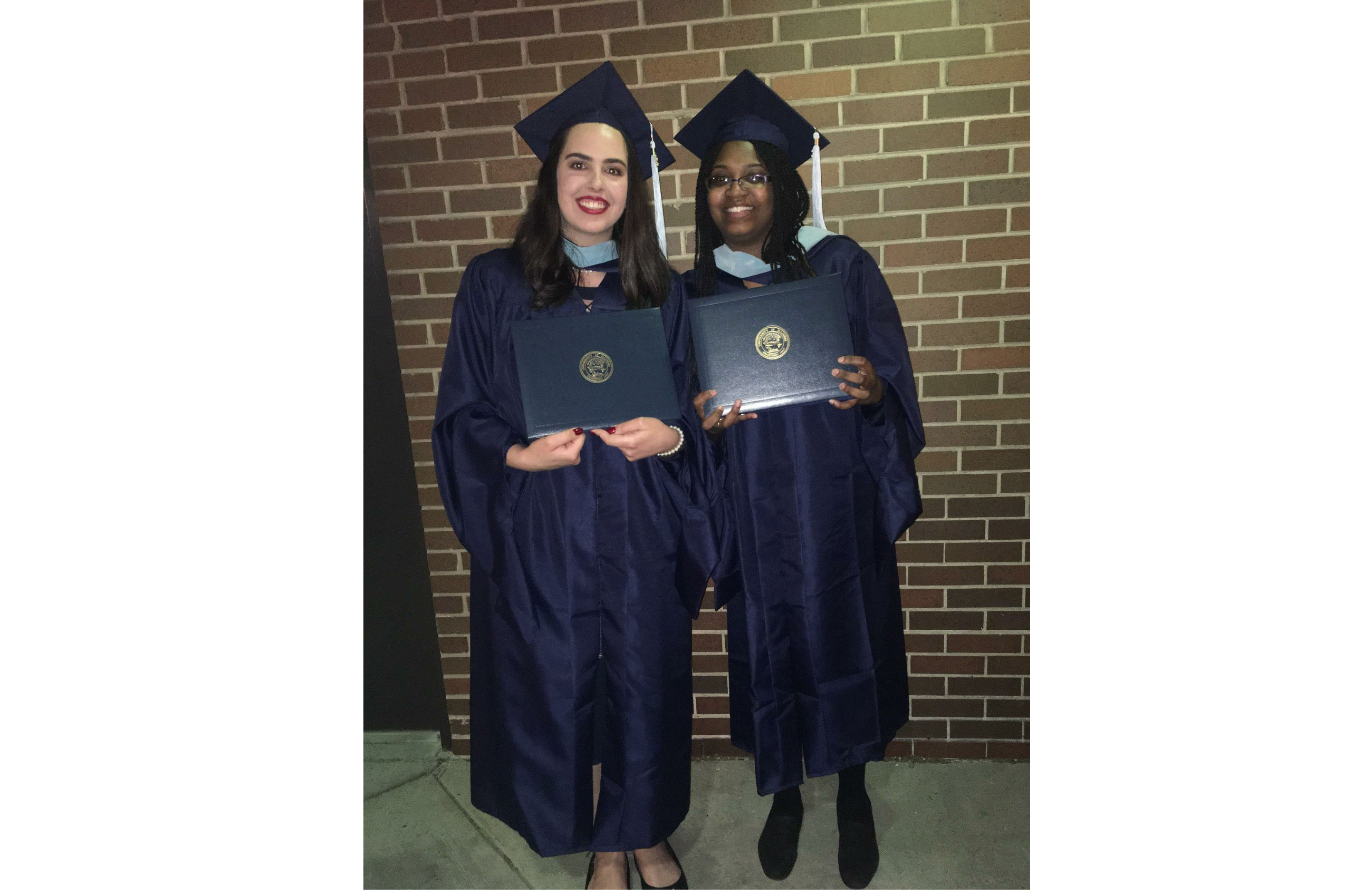 two young women stand side-by-side, wearing graduation caps and gowns, holding their diplomas