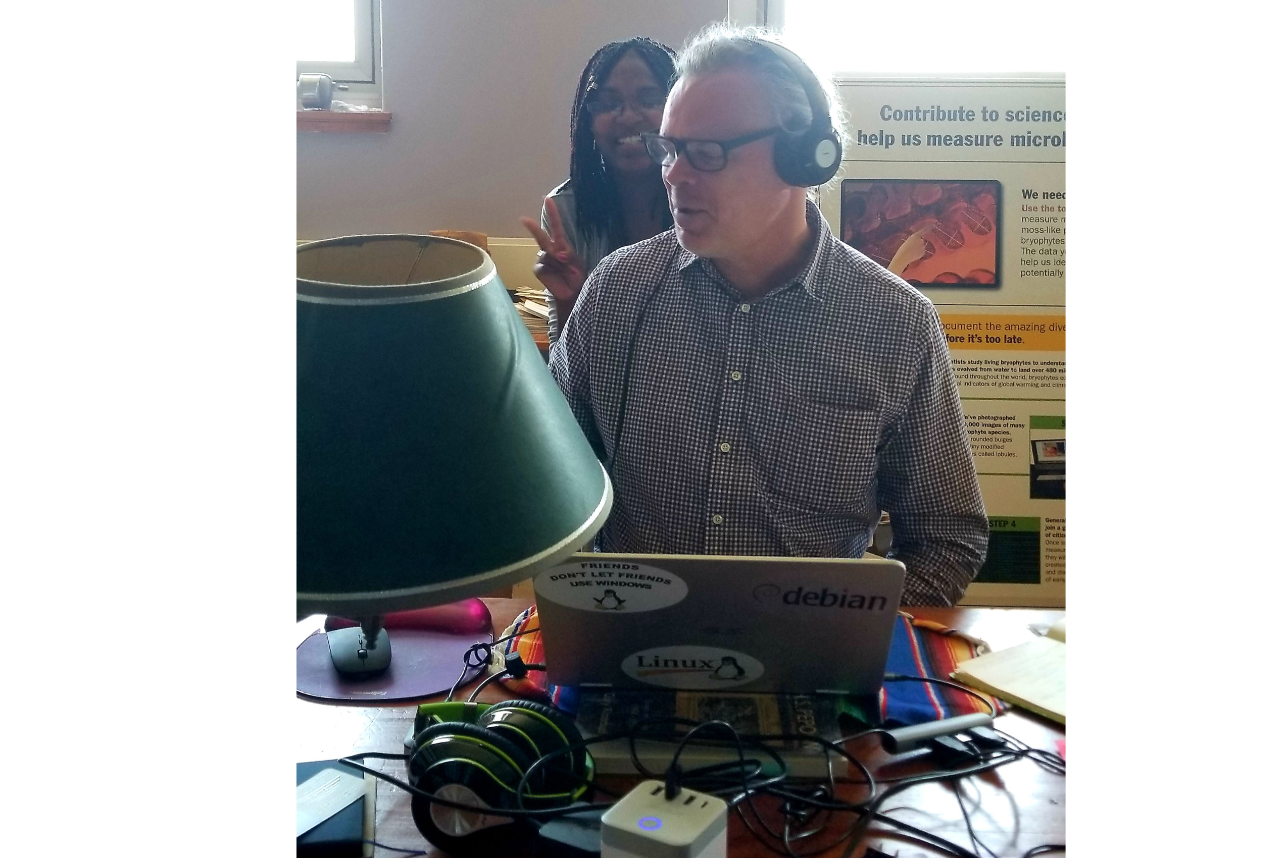 A man wearing glasses and headphones sits working on a laptop. A young woman smiles looking over his shoulder.