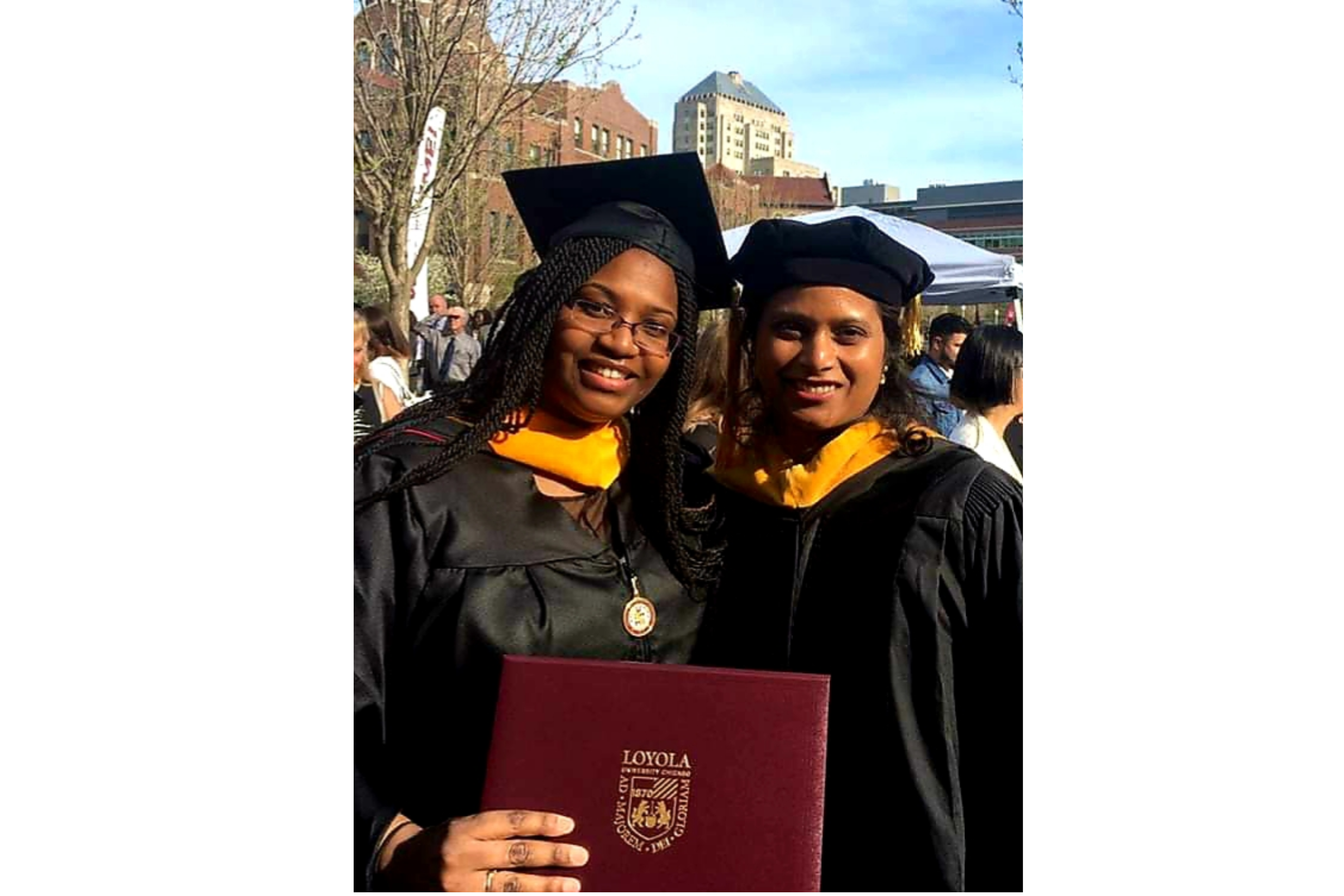 Two women wearing graduation regalia stand together, one holding a diploma.