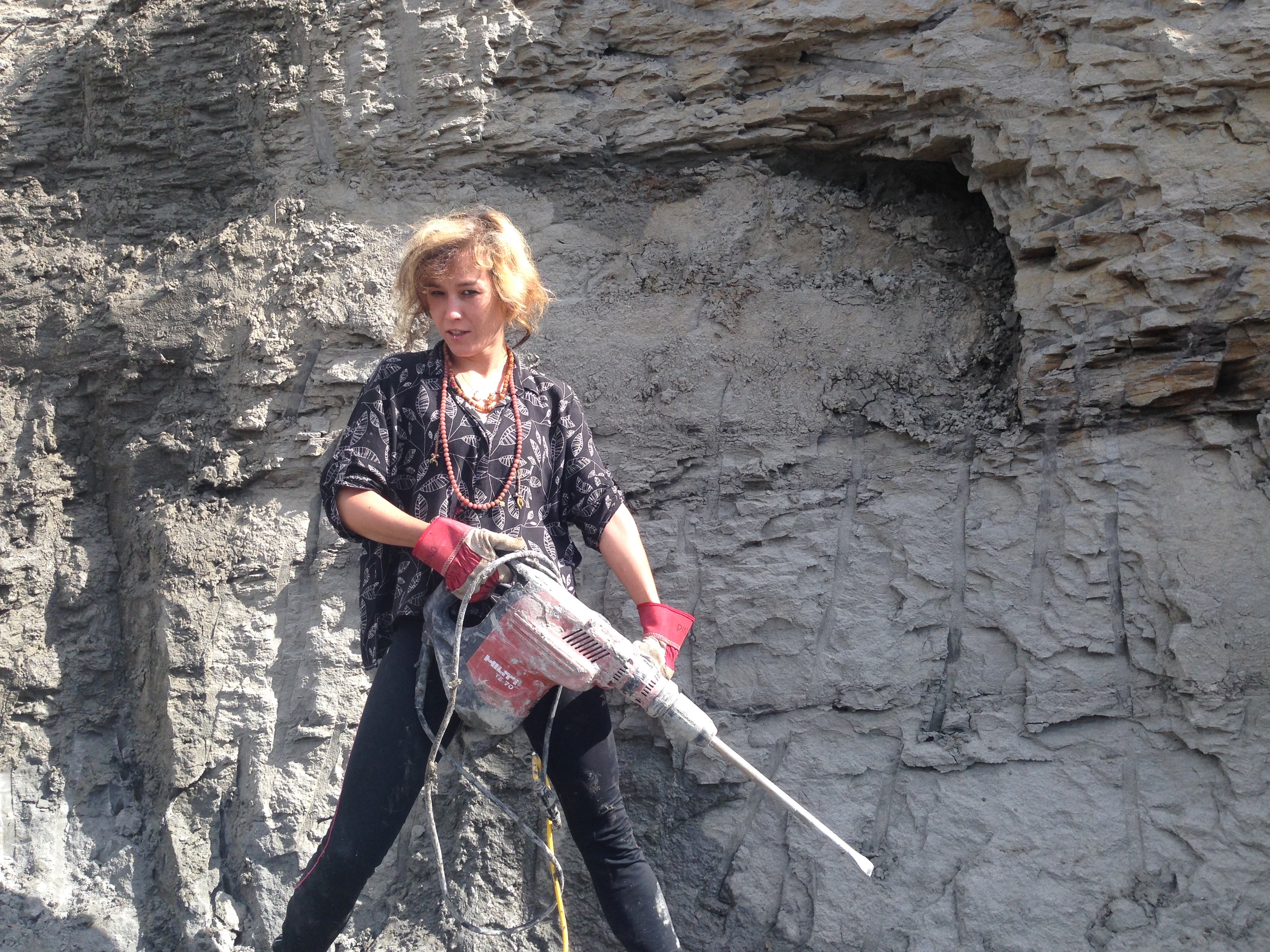 A woman holds a power drill, standing in front of a rock wall
