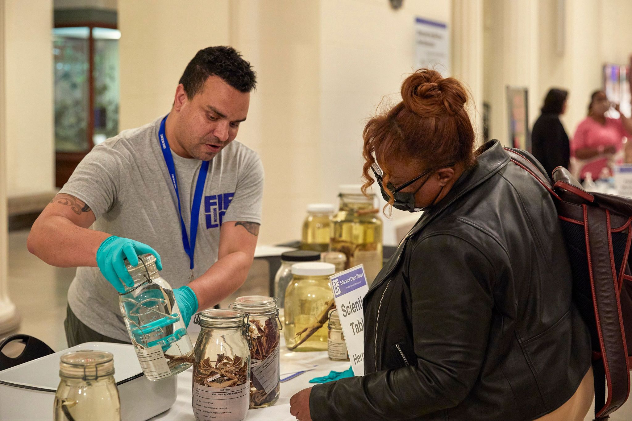 A museum staff member interacts with a visitor, showing specimens in jars.