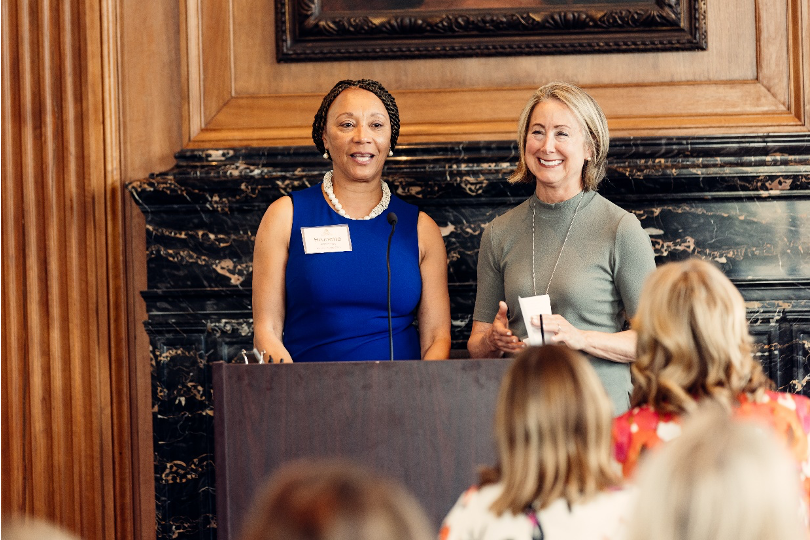 Two women stand at a podium presenting in a meeting room.