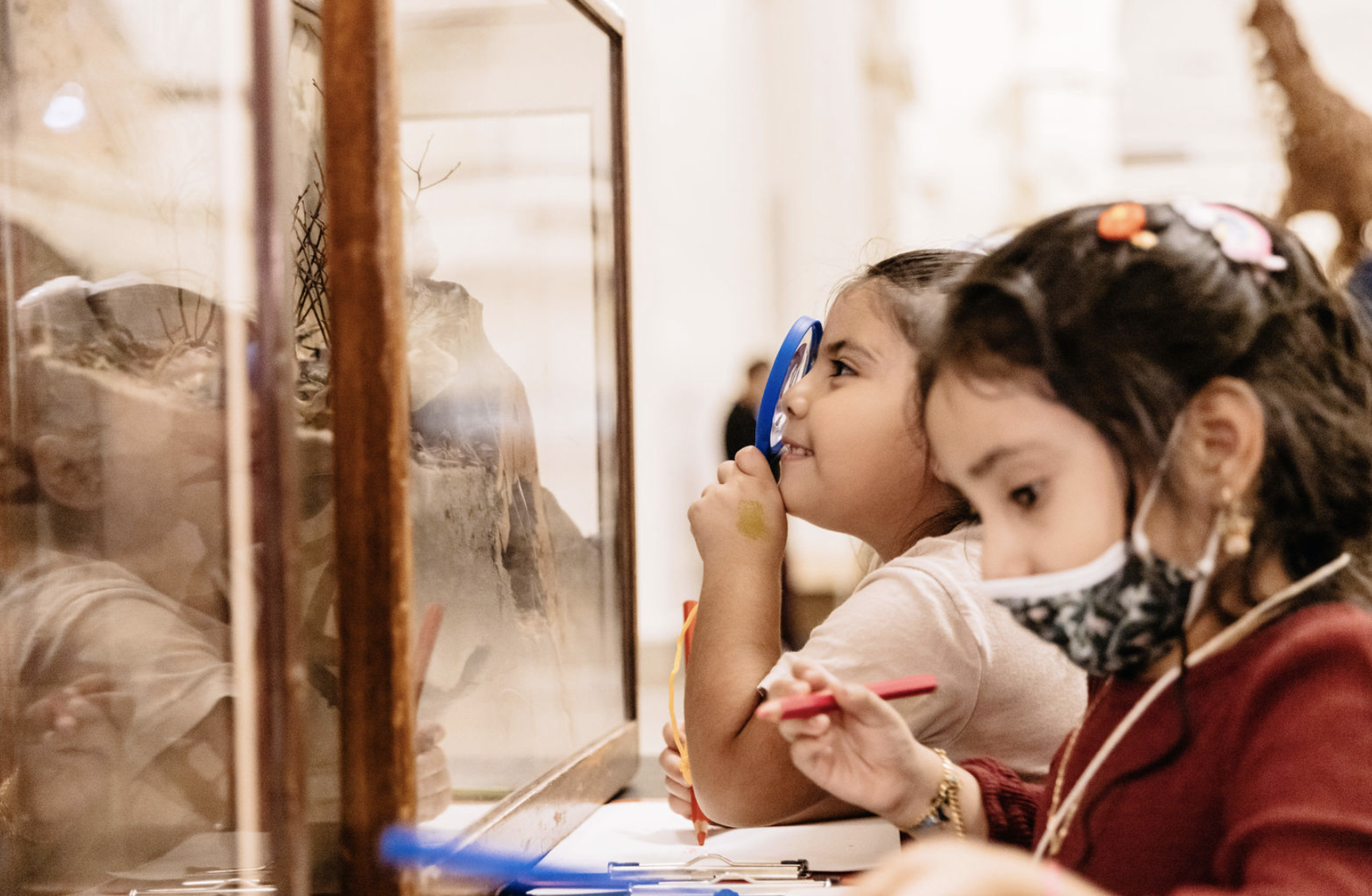 two children at a museum display case, one looking in with a magnifying glass, the other with a notebook and pencil.