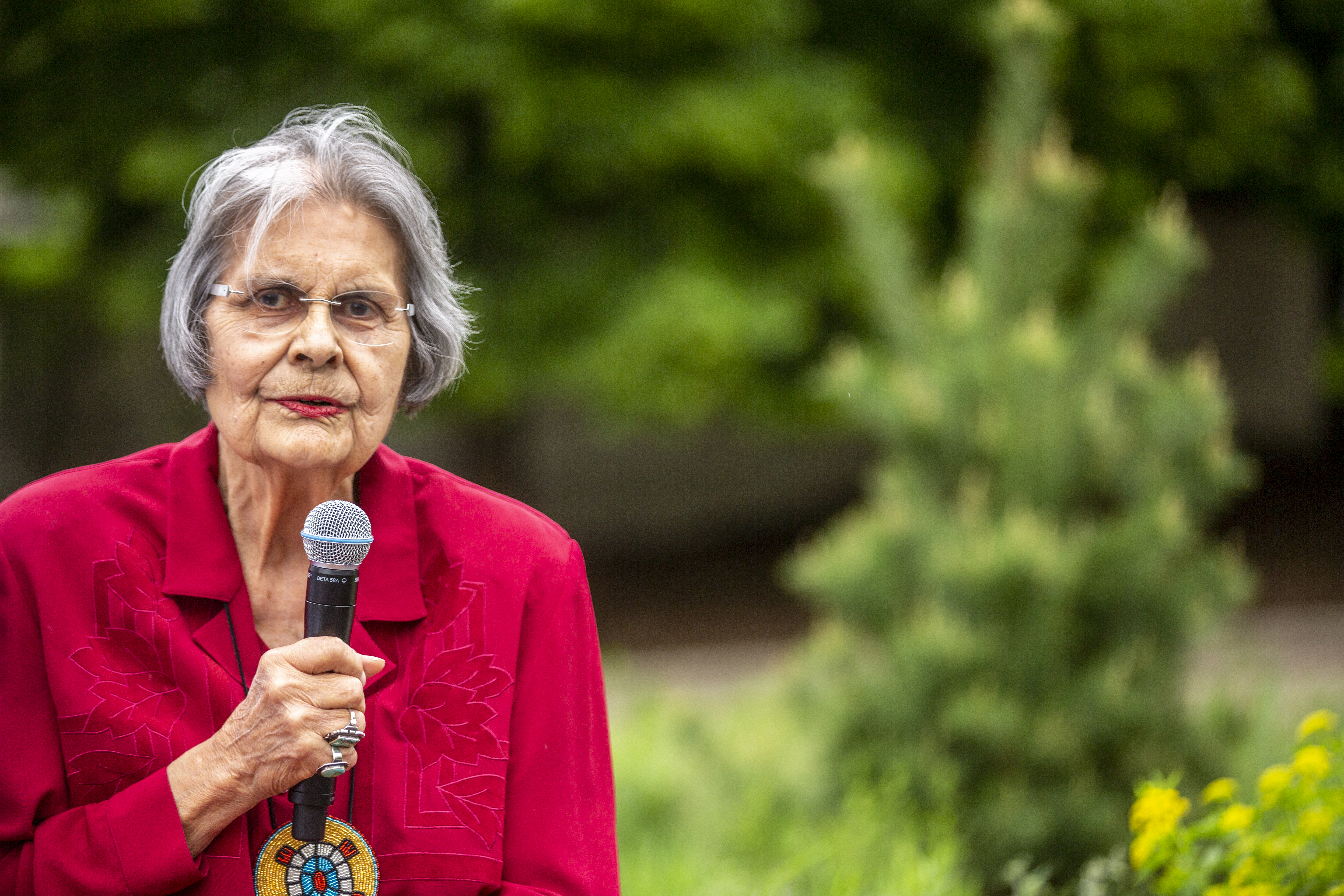 An woman with grey hair and glasses, wearing a red jacket and holding a microphone