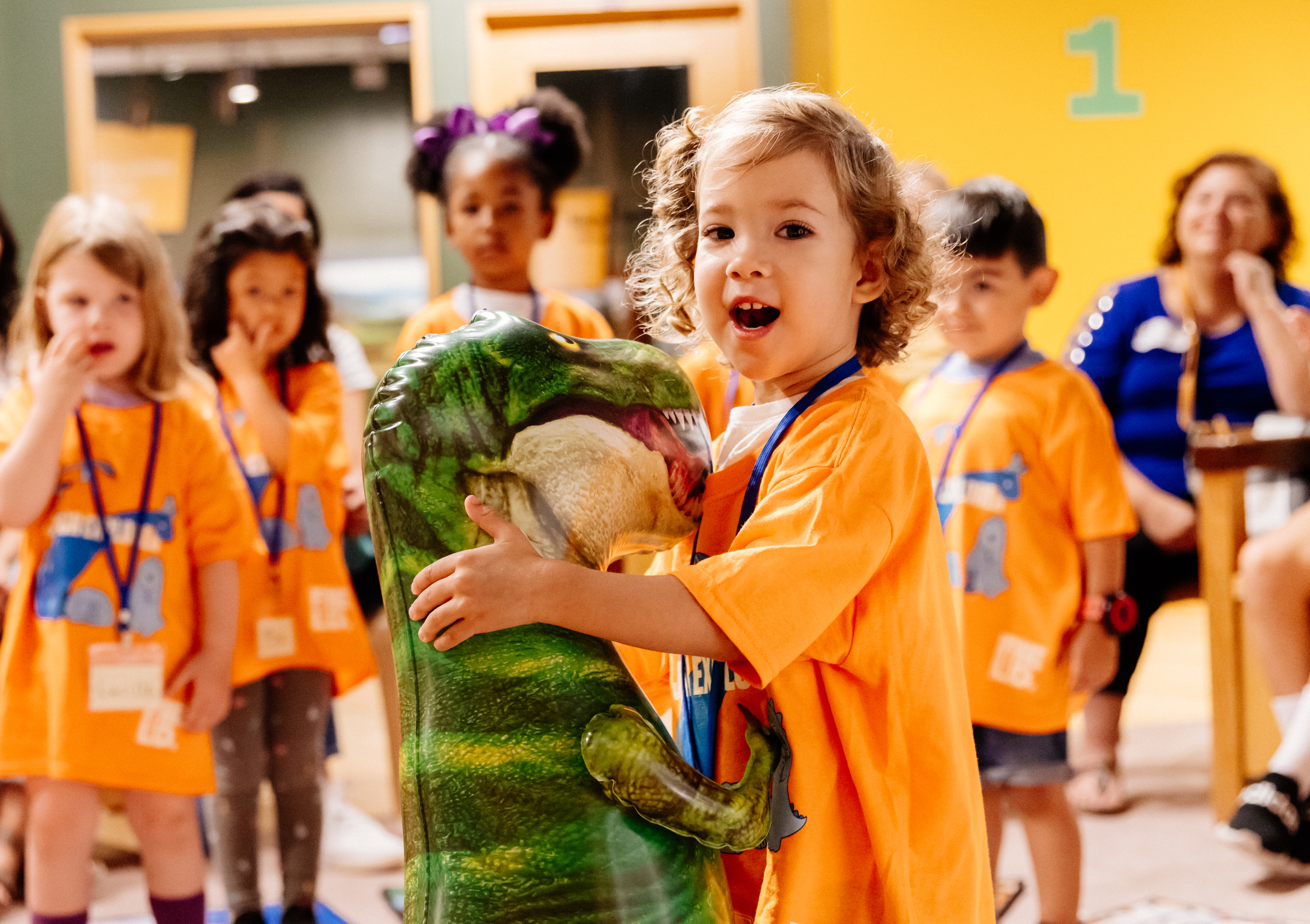 A young child holds a dinosaur balloon while a group of children can be seen in the background. All are wearing orange shirts
