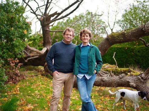 Two people stand side-by-side in front of a large fallen tree. A dog stands near them.