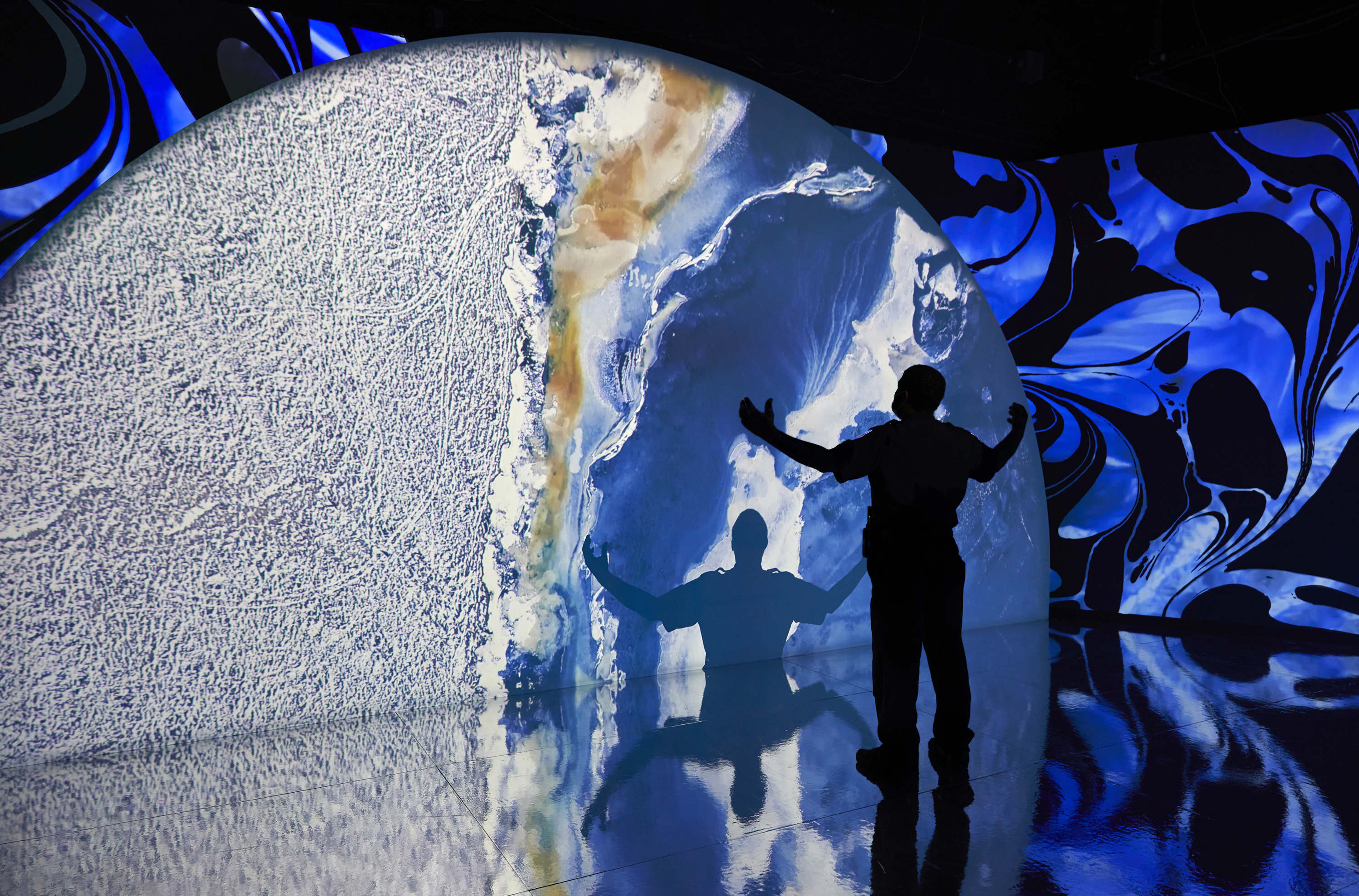 A man in silouette, stands with his arms outstretched in a museum exhibition. His shadow is cast onto a multi-media wall panel and the smooth, shiny floor.